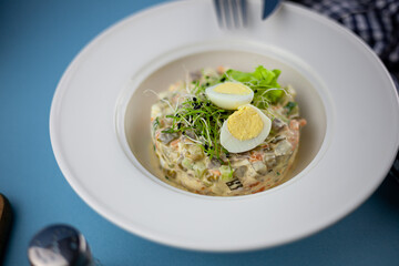 Traditional salad Olivier. Christmas and New year food. White plat with fork and knife on blue background with copy space. Top view, flat lay. Microgreen, quail eggs. Serving at a restaurant.