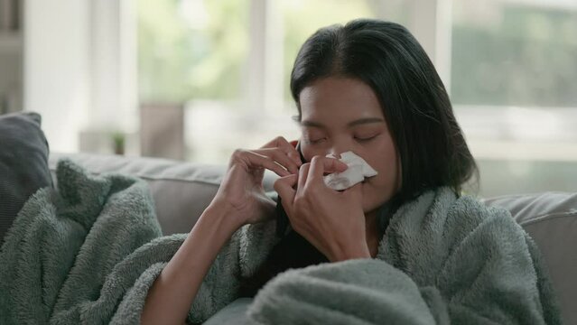 Footage of unhappy sad Asian woman in cloak suffering from fever and flu on sofa while talking with mobile phone in the living room. Health care and medical concepts.