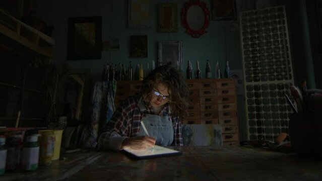 Graphic Designer Using Tablet Computer At Night. 30s woman drawing on tablet sitting in workshop in dark.