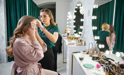 Female stylist in sterile gloves holding client hair lock and pulling string while working in visage studio. Woman sitting at dressing table and touching temple while having beauty procedure in salon.