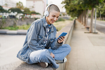 Young bald woman listening music playlist while using mobile phone outdoor - Focus on face