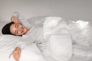 Glad young european lady sleeping on white bed with closed eyes, waking up, stretching body,...