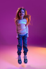 Happy, positive kid. Portrait of little girl, child in casual clothes posing, cheerfully jumping over purple studio background in neon light. Concept of childhood, emotions, fashion, lifestyle