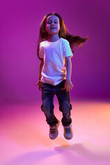 Having fun. Portrait of little kid, child in white T-shirt posing, cheerfully jumping over purple background in neon light. Concept of childhood, emotions, fashion, lifestyle