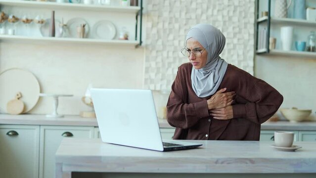 Young Muslim woman with severe heartache feels bad A female in a hijab suffers from chest pain while sitting at home working on a laptop computer alone indoor having heart attack disease