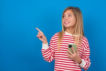 Wow!! excited caucasian teenager girl wearing striped T-shirt over blue background showing mobile phone with open hand gesture