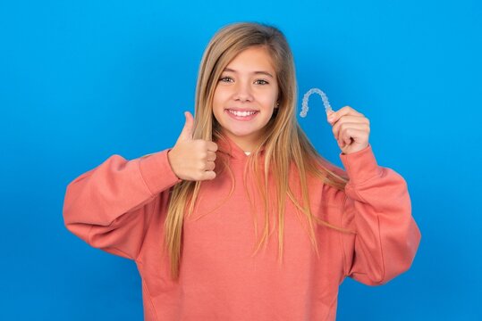 caucasian teenager girl wearing pink sweater over blue background holding an invisible braces aligner and rising thumb up, recommending this new treatment. Dental healthcare concept.