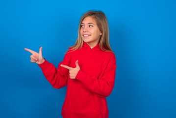 Optimistic caucasian teenager girl wearing red sweater over blue background points with both hands and  looking at empty space.