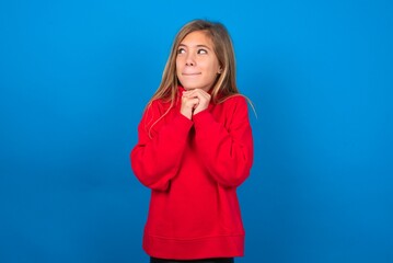 Curious caucasian teenager girl wearing red sweater over blue background keeps hands under chin bites lips and looks with interest aside.