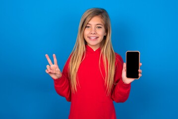 caucasian teenager girl wearing red sweater over blue background holding modern device showing v-sign