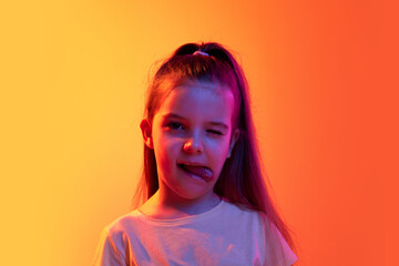 Playful kid. Portrait of little girl, child posing with funny face over gradient orange background in neon light. Concept of childhood, emotions, fashion, lifestyle, facial expression