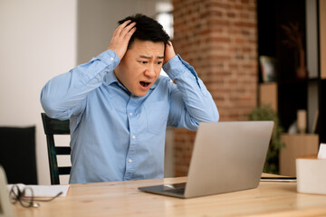 Shocked asian businessman looking at laptop screen and touching head, reading shocking news, working remotely at home