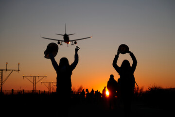 Fototapeta na wymiar Two backlit women raise their arms to airplanes at sunset as a sign of travel and adventure.