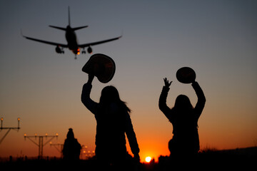 Obraz na płótnie Canvas Two backlit women raise their arms to airplanes at sunset as a sign of travel and adventure.