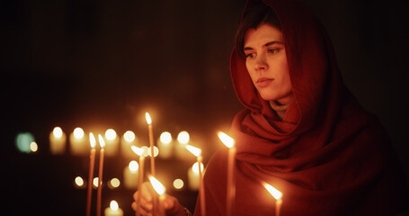 Aesthetic Shot of Young Christian Woman with a Head Scarf Lighting a Candle in Church, Praying and...