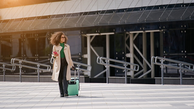Young african woman walking outdoors carrying a suitcase and going to travel by airplane at modern airport. Vacations, travel and active lifestyle concept    