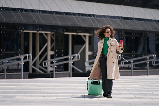 Young african woman walking outdoors carrying a suitcase and going to travel by airplane at modern airport. Vacations, travel and active lifestyle concept