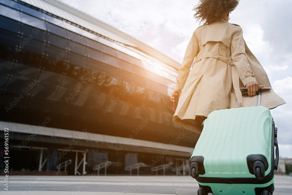 Wall mural Young african woman walking outdoors carrying a suitcase and going to travel by airplane at modern airport. Vacations, travel and active lifestyle concept    