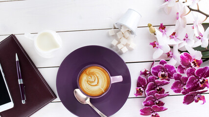 cup of cappuccino coffee on violet plate, diary, rich pen, phone and orchid flower on white colored wooden table, top view