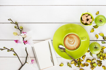 cappuccino coffee in green cup,  orchid flower, notebook and silver pen,  green dry flower decor scattered on white painted wooden table, top view - 569162916