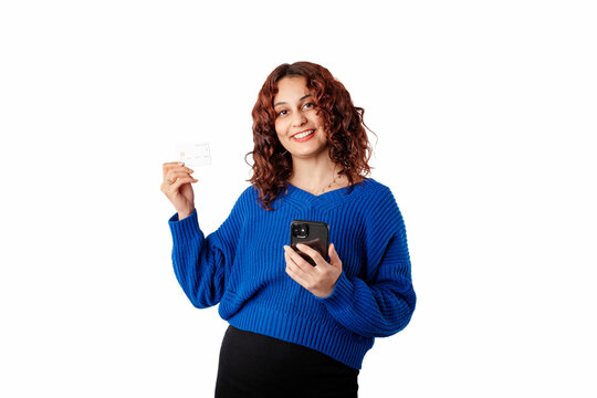 Portrait Of Woman Standing Isolated Over White Background Holding A Smart Phone And Looks At The Camera Smiling And Showing Credit Card. Recommends Effortless, Safe, Online Shopping.