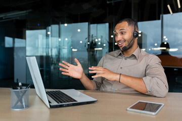 Young successful African American man with video call headset working inside office with laptop, businessman in shirt talking to client advising consumer online, helping with setting up device.
