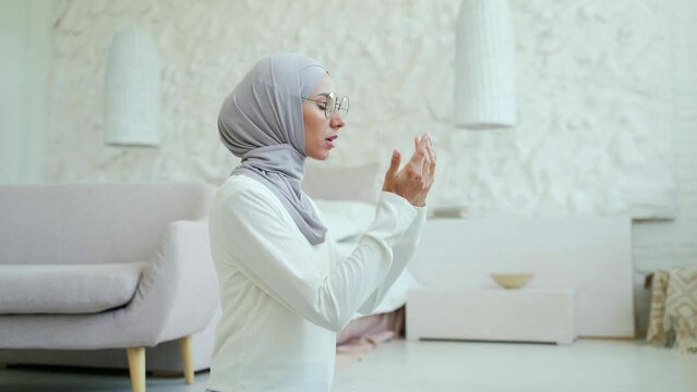 A young Muslim woman in a hijab prays at home. Alone in a bright living room in the house worshiping to the GOD after reading Quran while wearing veil in the lounge Eastern culture religion, lifestyle