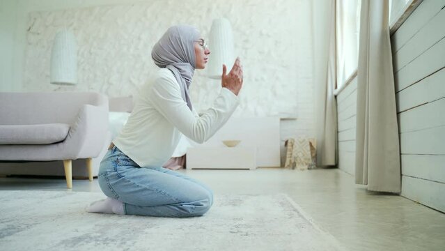 A young Muslim woman in a hijab prays at home. Alone in a bright living room in the house worshiping to the GOD after reading Quran while wearing veil in the lounge Eastern culture religion, lifestyle