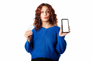 Fototapeta premium Cheerful woman wearing blue pullover sweater isolated over white background showing credit card and the blank white mockup screen of the phone, looks at the camera with a puzzled expression. Wow mouth