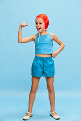 Showing muscles. Having fun. Portrait of little girl, child in swimming cap posing over blue studio background. Concept of childhood, emotions, fun, fashion, lifestyle, facial expression
