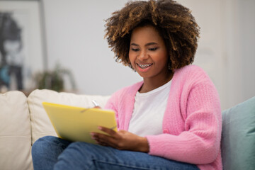 Positive millennial black woman using notepad and pen at home