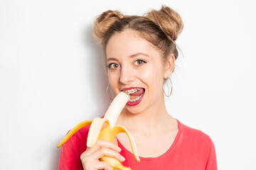 Young caucasian woman with two hair bundles wearing braces eating banana and enjoying it  - obrazy, fototapety, plakaty