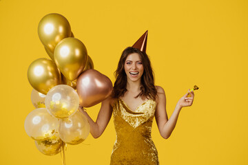 Overjoyed lady with festive balloons, birthday cap and party blower celebrating special occasion over yellow background