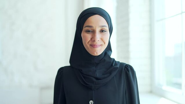 closeup portrait of a pretty young Muslim woman in a black hijab standing and smiling indoors at home Headshot of an Islamic happy female looking at the camera on a light office room close up face