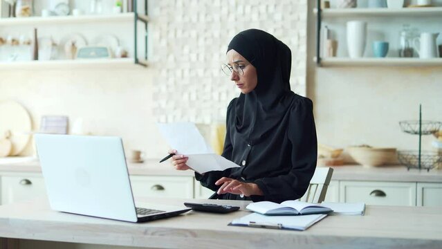 young Muslim woman works from home online, calculates family finances or will study remotely Female in hijab uses laptop computer notes Business woman uses app or website manages personal finances