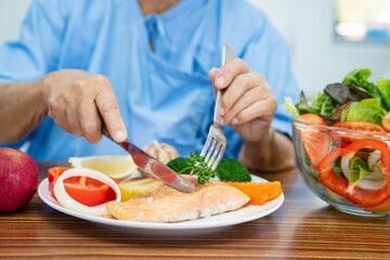 Asian senior or elderly old lady woman patient eating Salmon steak breakfast with vegetable healthy food while sitting and hungry on bed in hospital.