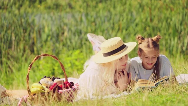 Cheerful woman and daughter lying on blanket and reading, picnic by the lake
