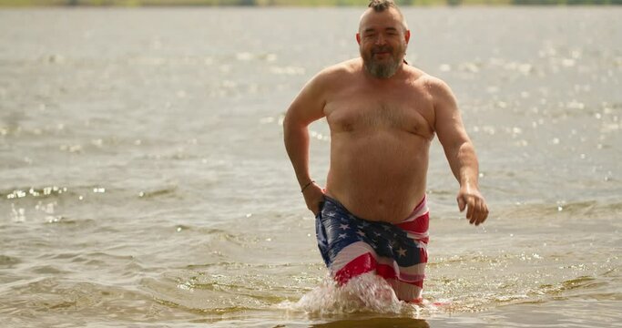 A bearded man, after swimming in the river, wrapped himself in a towel with a painted american flag on it. Swimming in the water. Journey to the lake.
