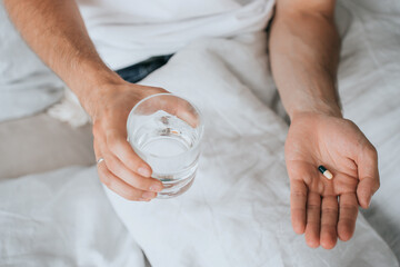 Cropped image of mans hands holding glass of water and pill sitting on bed, gets ill. Treatment, medicine, pharmacology. Sick guy holds drug, feels bad. Pandemic, virus. Recovery, illness.