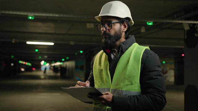 Indian contractor mechanic in helmet and glasses writing in paper examination parking lot write in clipboard. Arabian bearded man worker of manufacture work engineer in hard hat daily check inspect