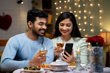 Happy couple using mobile phone while having drinks during romantic candlelight dinner - concept of...