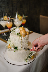 Bride and a groom is cutting their rustic wedding cake on wedding banquet. Hands cut the cake with delicate white flowers and golden decor.