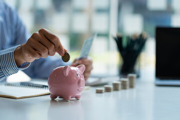 Hands of a young Asian businessman Man putting coins into piggy bank and holding money side by side...