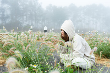 Asian woman wearing hoodie with thick long-sleeved hat, Protection against cold, foggy mornings, She was sitting on ground surrounded by flowers that grew naturally, sit look at flowers warmly