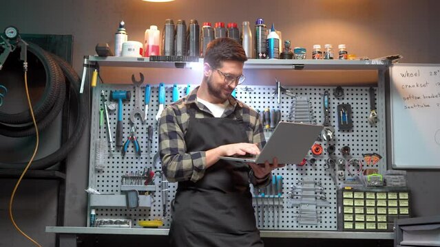 Young adult caucasian male craftsman holding and using laptop online typing standing at modern garage with tools. Attractive handsome happy man mechanic wearing glasses and apron.