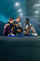 Fitness gym instructor showing workout plan on tablet to two fit woman in sportswear with towel. They are sitting on the gym floor, resting after workout.