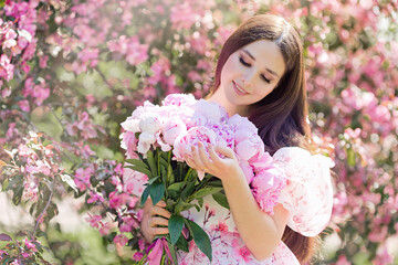 A happy brunette in a dress, with a large bouquet of peonies, stands near pink blooming apple trees