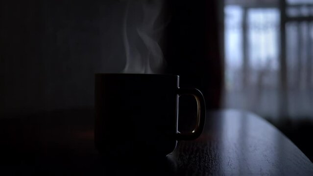 Close-up of a cup of hot tea stands on a table in a dark room. Fragrant steam rises over a cup of tea. Hot drink for breakfast in the early morning. Space for copy paste lettering or logo.