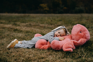 Little child wearing a bunny costume, laying on a big pink plush bunny toy, in an open field.