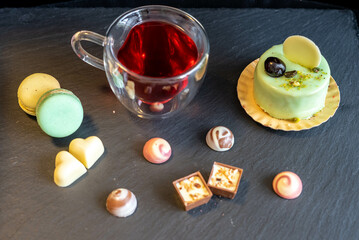 Pistachio cake with macaroons, chocolate pralines and red tea in a heart shaped glass mug on a black background slate
